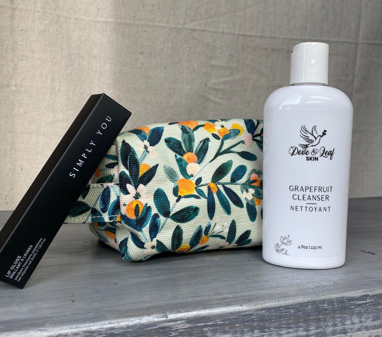 Cosmetic bag with floral design,Simply You Mascara, and white bottle facial cleanser  on a wooden surface.