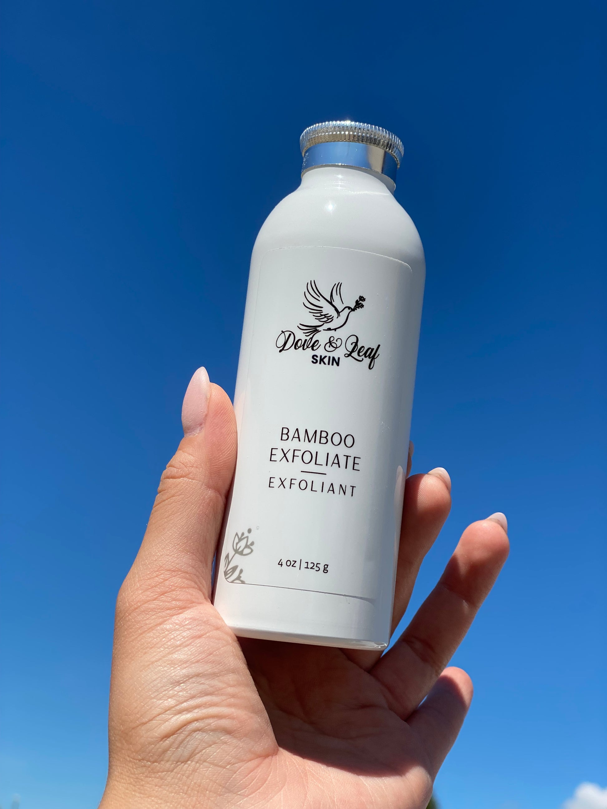 Hand holding a bottle of 'dove & leafl' bamboo exfoliant against a blue sky.